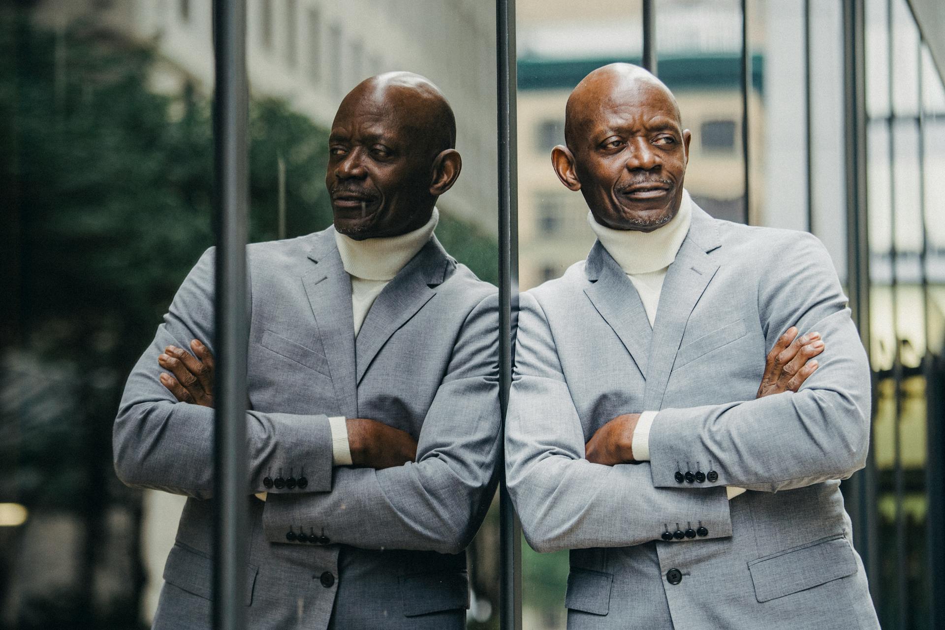 Elegant man in a suit poses confidently next to a glass wall outdoors, showcasing his reflection.