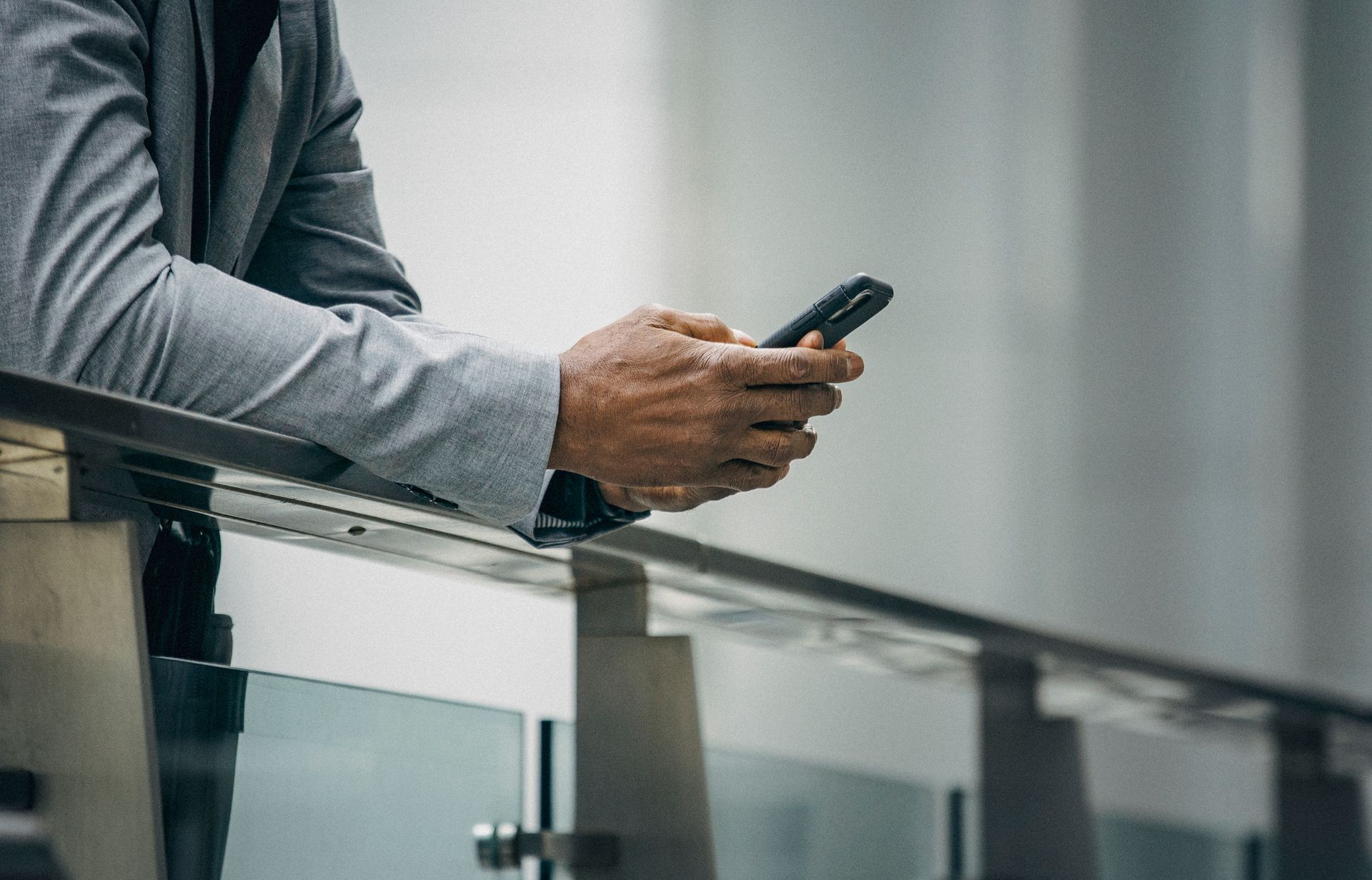 Crop anonymous black businessman in formal suit leaning on fence and using phone in soft daylight