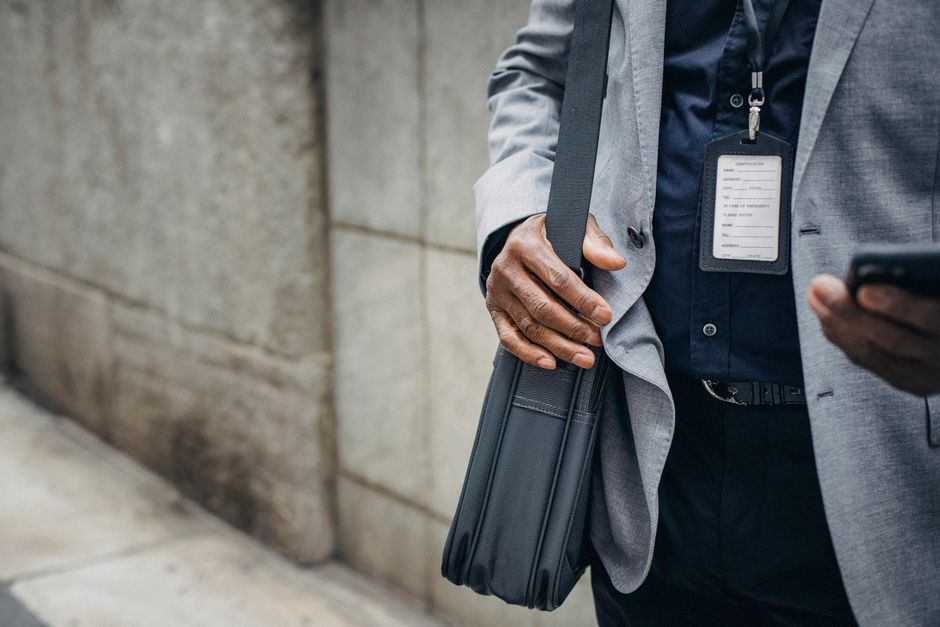 Crop anonymous African American entrepreneur in gray jacket with satchel and pass card browsing internet on smartphone on pavement near stone wall in city in daylight Crop anonymous African American entrepreneur in gray jacket with satchel and pass card browsing internet on smartphone on pavement near stone wall in city in daylight