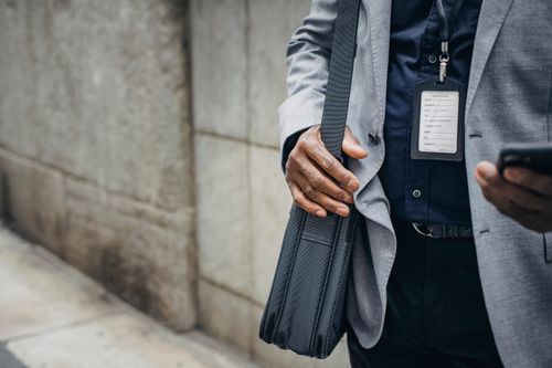 Free Crop anonymous African American entrepreneur in gray jacket with satchel and pass card browsing internet on smartphone on pavement near stone wall in city in daylight Stock Photo