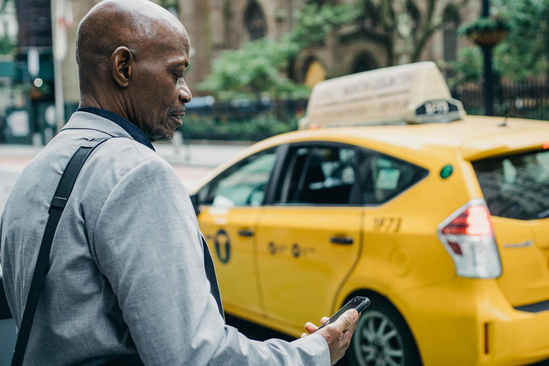 Businessman checks his phone while standing next to a yellow New York taxi on a city street.