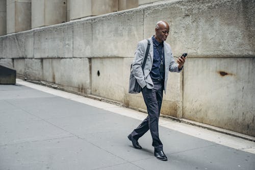 Full body content African American male in classy formal suit walking with hand in pocket along city sidewalk and browsing mobile phone