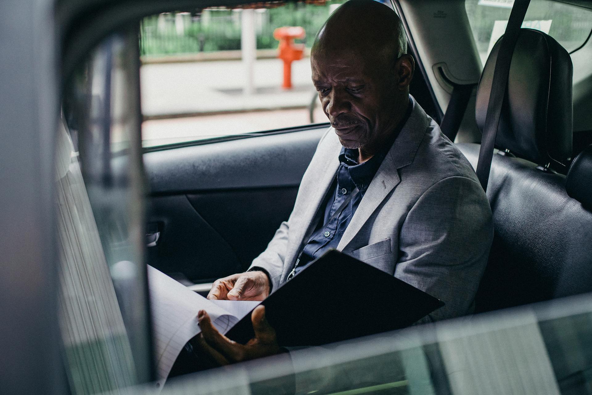 Side view serious African American businessman in formal clothes sitting in car back seat and reading documents