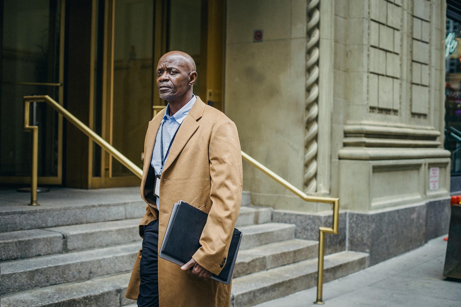 Pensive African American executive manager with important documents looking away and walking on street near gray building