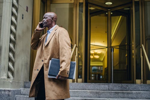 African American male in formal outfit talking on mobile phone near entrance of modern building