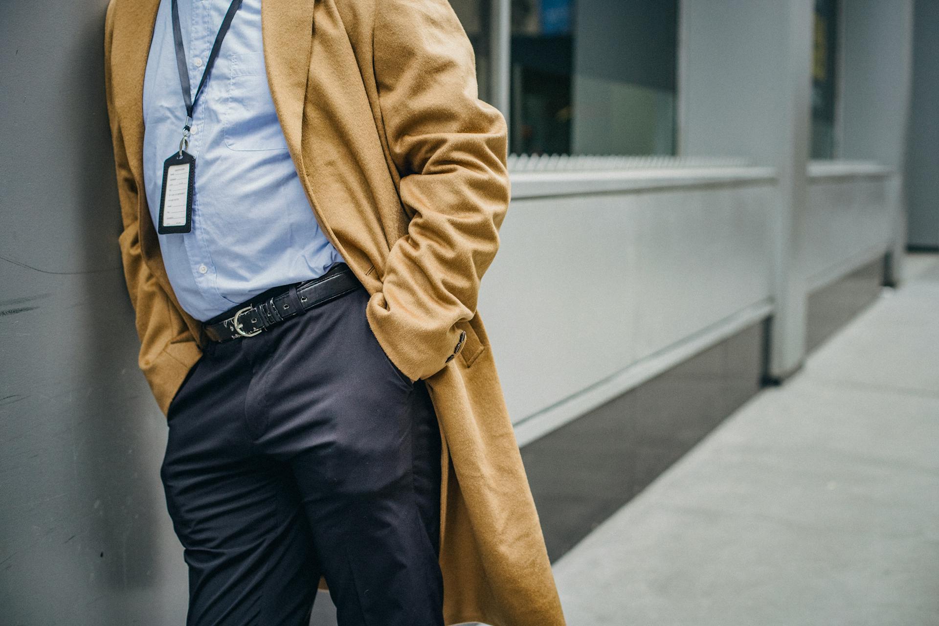 A well-dressed man in an overcoat leans against a wall in an urban setting, exuding professionalism and style.