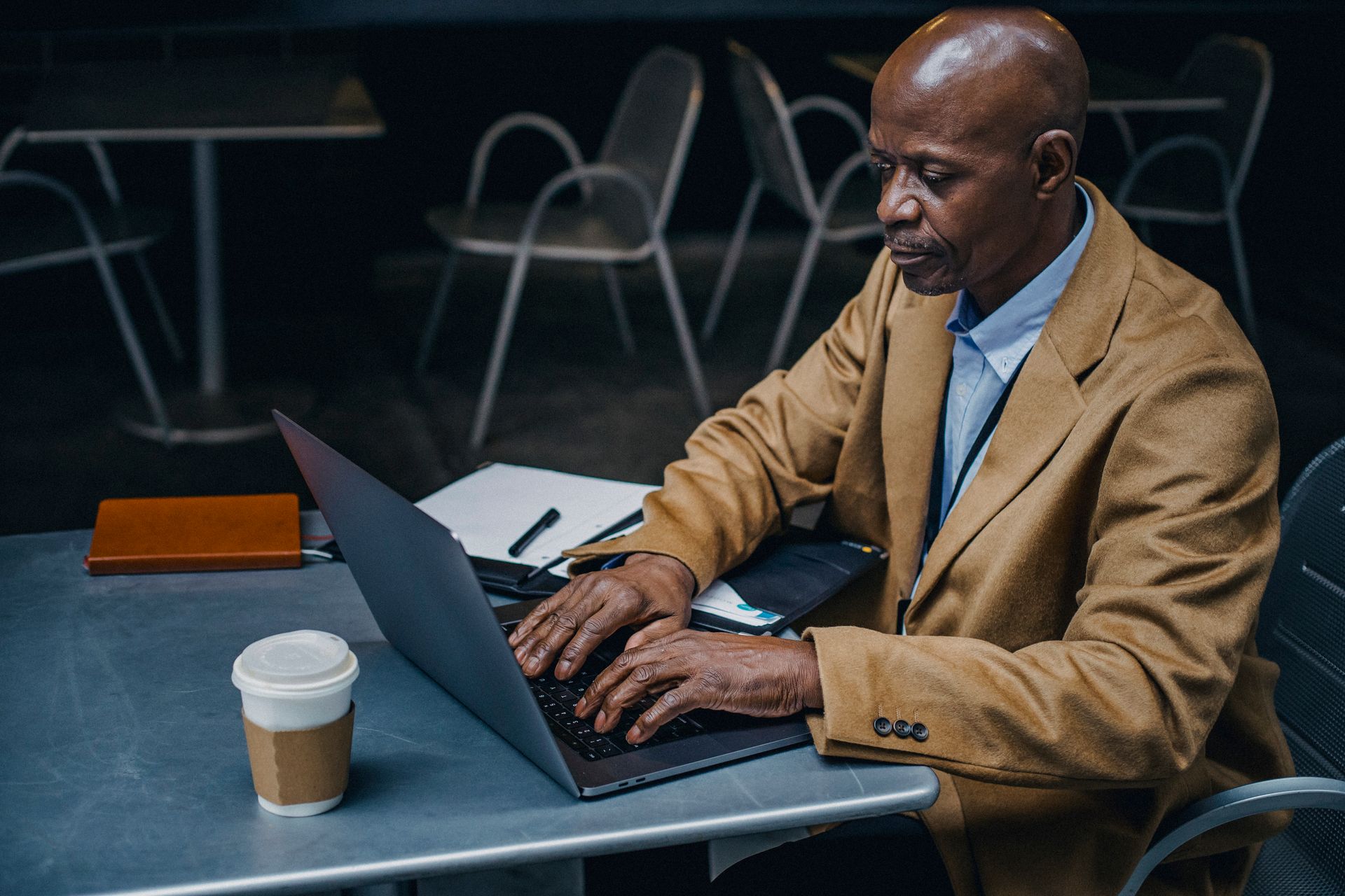Confident man in a coat working intently on a laptop indoors, with a coffee cup beside him.