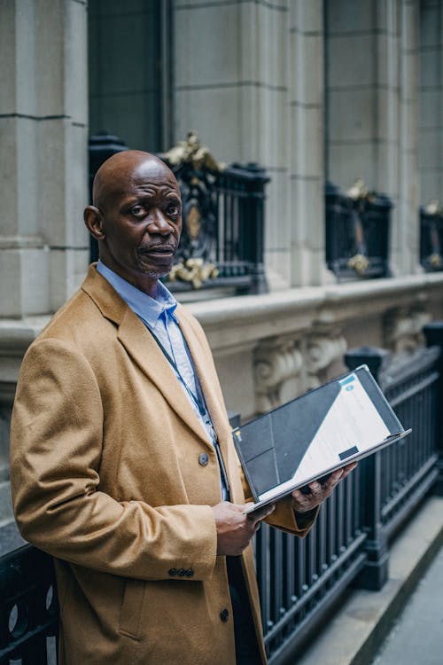 Middle aged African American male in elegant outfit looking at camera and reading report on street near building