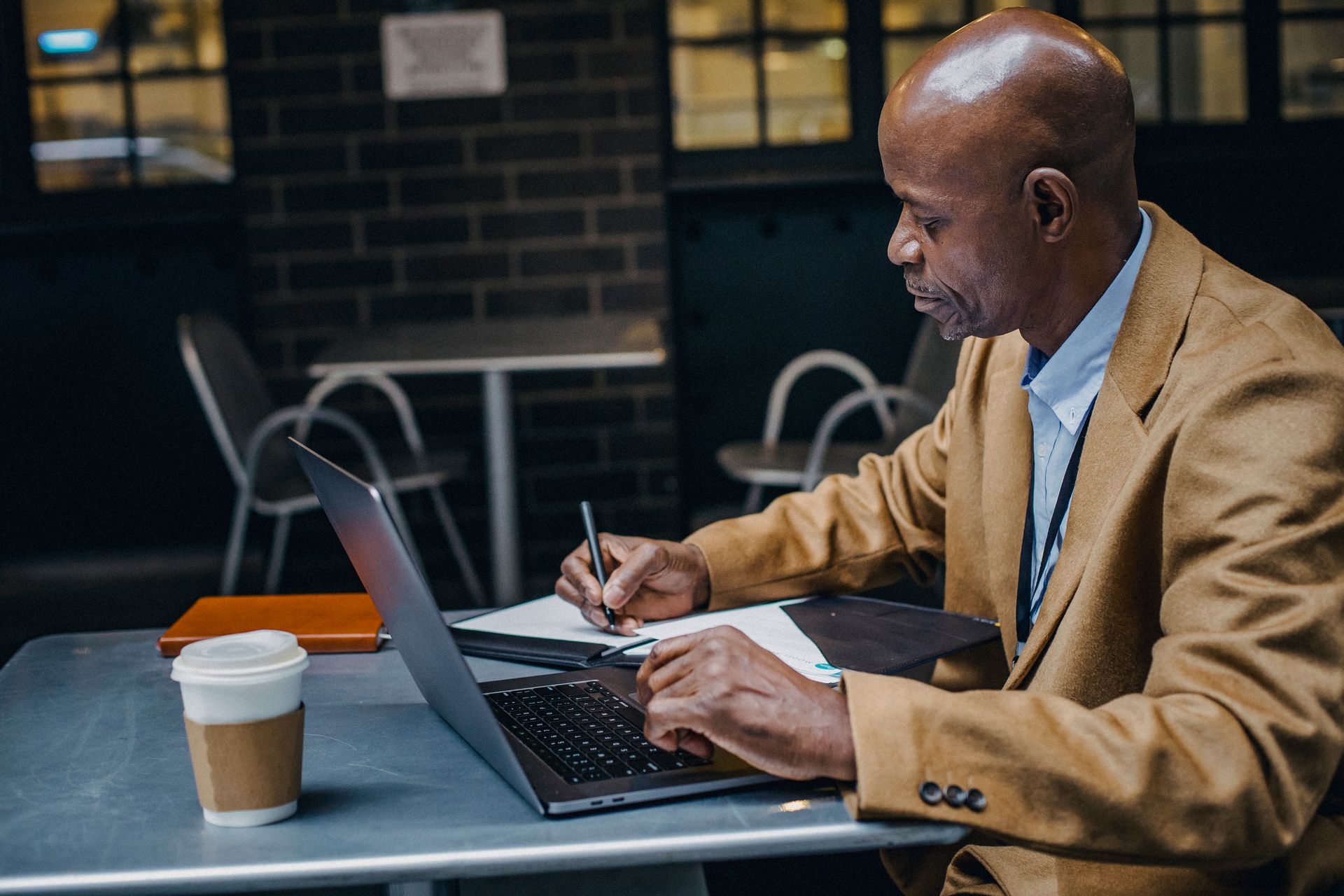 Side view of focused African American freelancer sitting at table with laptop and making notes on paper in cafe