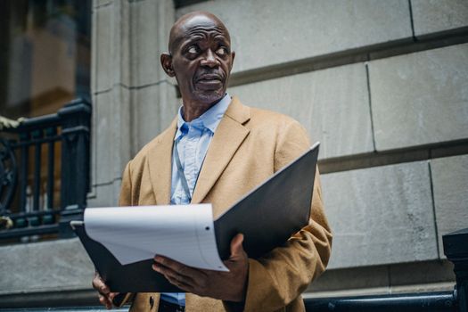 Low angle of gloomy African American man in trendy outfit standing with folder with documents in hands and looking away in daytime