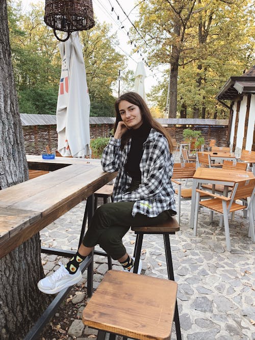 Positive young woman in stylish outfit relaxing on stool at wooden counter in cozy outdoor cafe on autumn day