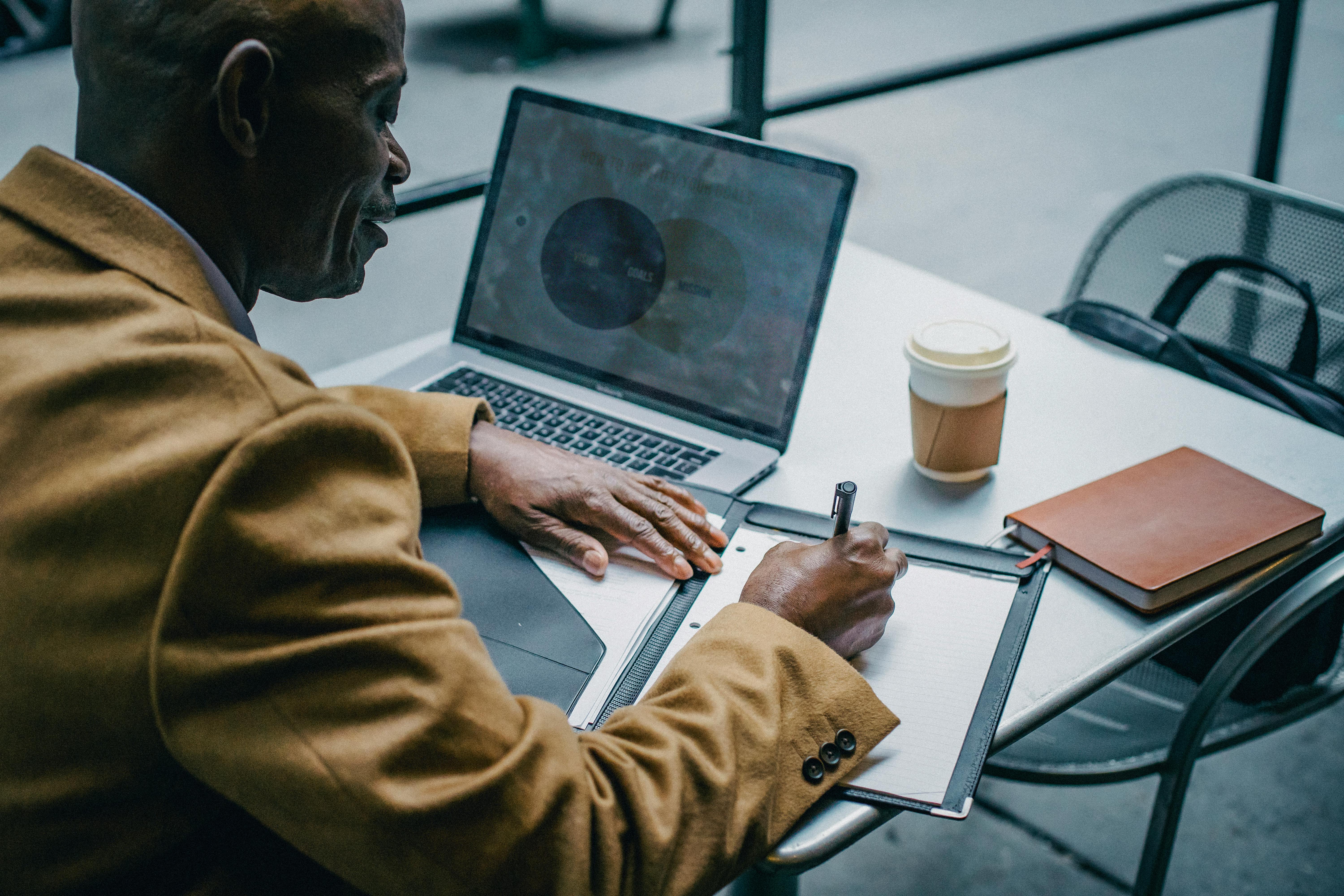 crop black businessman writing on paper near laptop in cafeteria