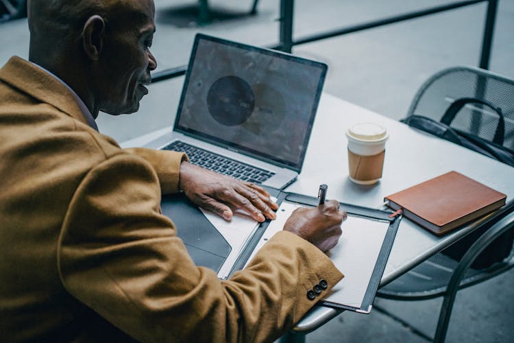 Crop Black Businessman Writing On Paper Near Laptop In Cafeteria