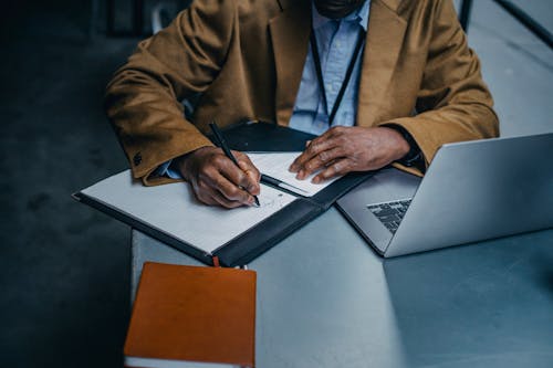 Crop black male executive writing on paper sheet near laptop