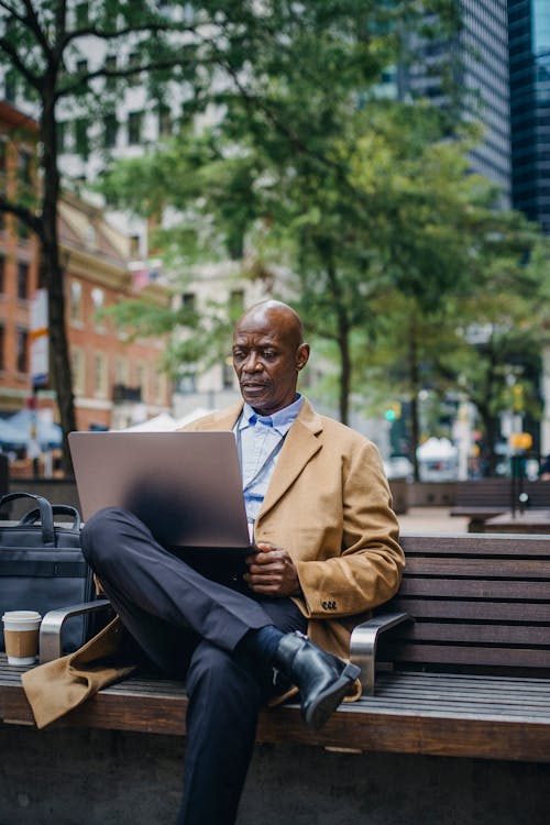 Focused African American male executive watching netbook while sitting with crossed legs on bench in town