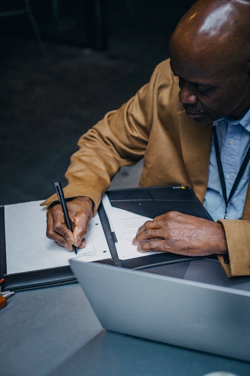 Crop focused African American male sitting at table with laptop and writing notes in planner