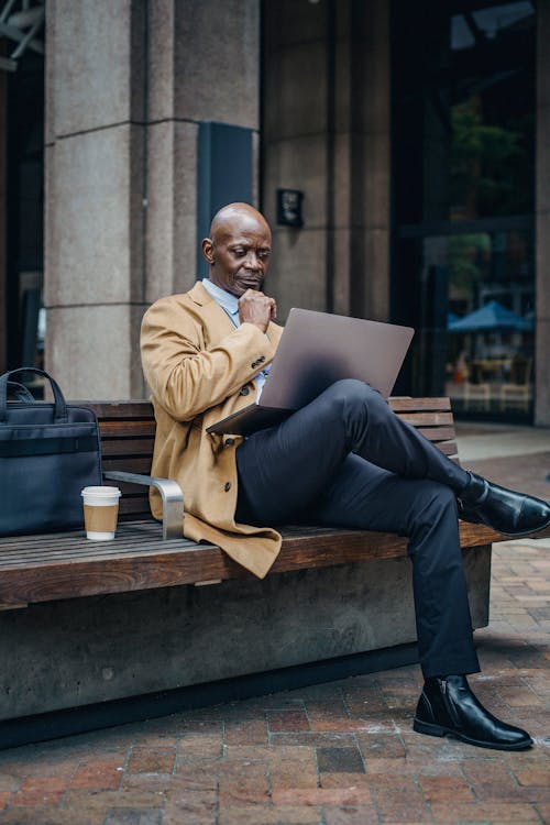 Pensive adult African American businessman wearing stylish formal clothes sitting on street bench and browsing netbook on laps while touching chin in thoughts