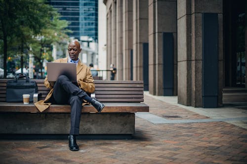 Free Full body pensive African American businessman in formal suit and coat sitting on street bench with legs crossed and browsing modern netbook in urban downtown Stock Photo