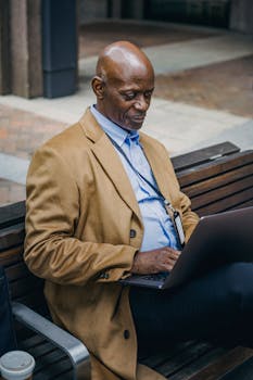 A well-dressed man sitting on a bench in the city, focused on his laptop, reflecting urban business life.