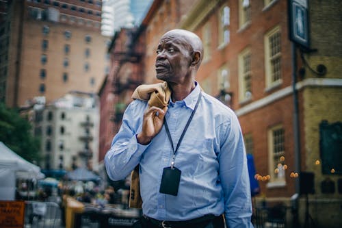 Serious adult African American businessman in formal clothes standing with jacket on shoulder on urban modern street and looking away thoughtfully