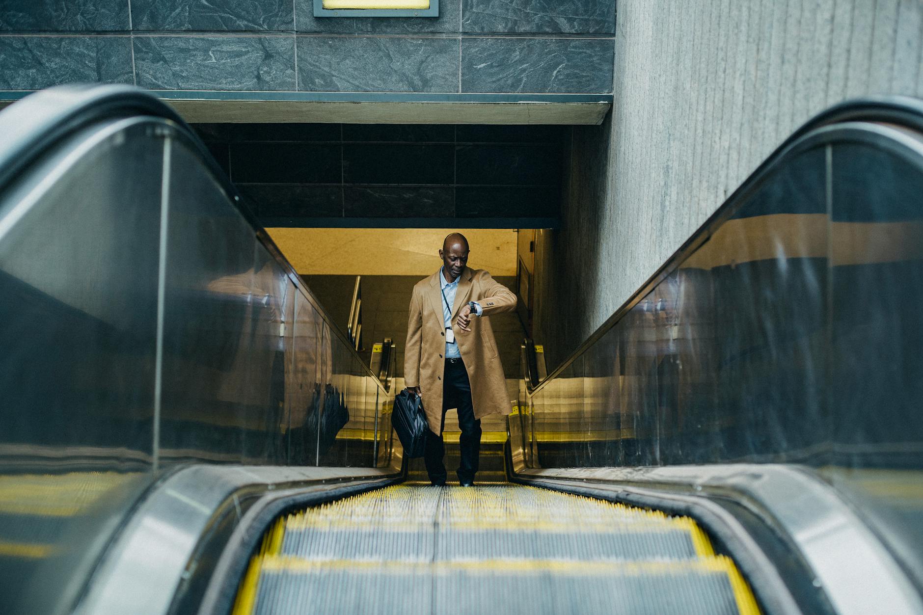 Contemplative black businessman riding escalator and looking at wristwatch