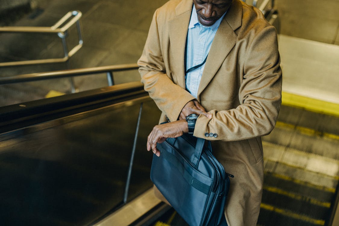From above crop punctual African American businessman riding escalator and checking time on wristwatch while leaving metro station