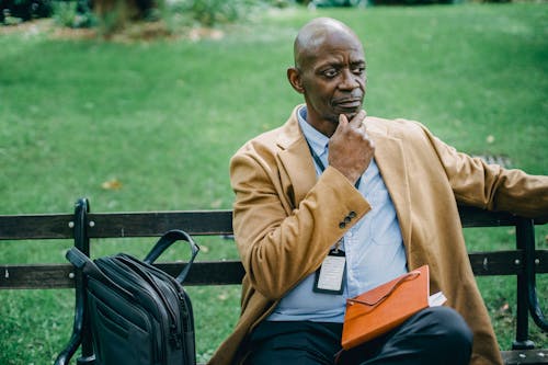 Contemplative African American male in trendy formal wear sitting on park bench with diary on laps and touching chin in deep thoughts