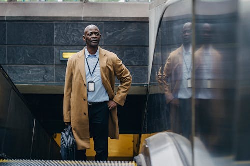 Confident adult African American businessman in formal clothes riding escalator with hand in pocket while leaving metro station and looking away thoughtfully