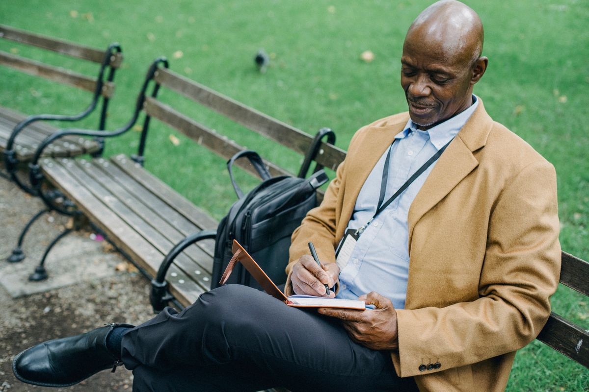 An older Black man in a tan blazer sits on a park bench taking notes in a notebook, focused and deliberate.