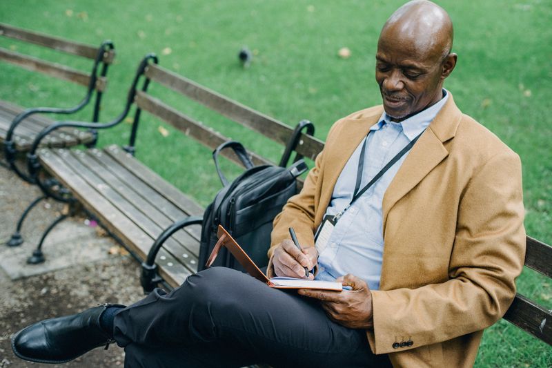 An older Black man in a tan blazer sits on a park bench taking notes in a notebook, focused and deliberate.