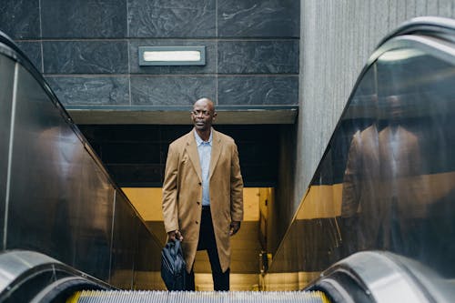 From above of mature well dressed black businessman in coat with briefcase standing on moving stairs while looking away in town