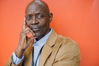 Confident middle aged bald African American male in elegant suit looking at camera with hand on cheek against orange background