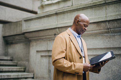 Side view of concentrated middle aged black male manager in formal clothes and coat standing near stairs of aged building and reading documents