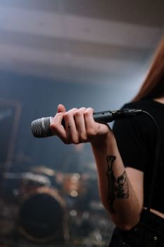 Close-up of a tattooed hand holding a microphone during a band rehearsal, with drums in the background.