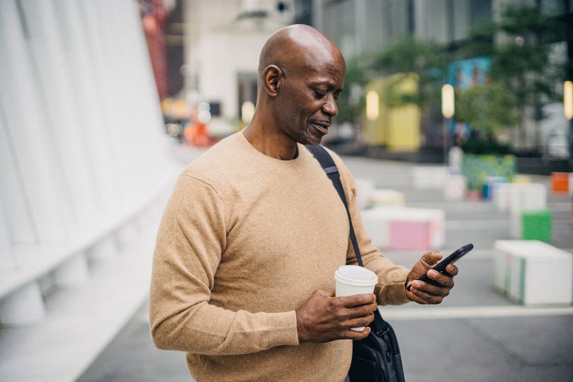 Confident middle aged African American man in stylish outfit standing on city street with takeaway coffee in hand and messaging on mobile phone