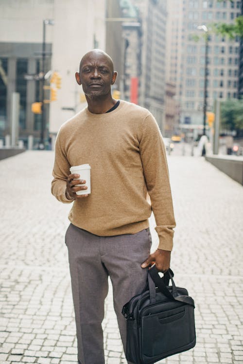 Free Confident stylish African American male freelancer standing on paved street in downtown with coffee to go and laptop bag in hands Stock Photo