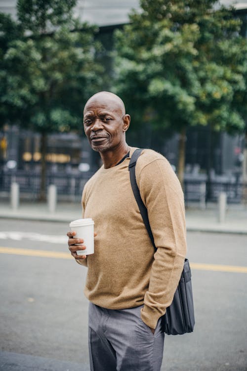 Side view of serious mature African American man in casual clothes drinking cup of takeaway coffee while standing on with hand in pocket and looking at camera