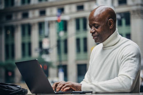 Side view of self employed concentrated mature bald black man in stylish turtleneck sitting in street cafe and typing on netbook during work