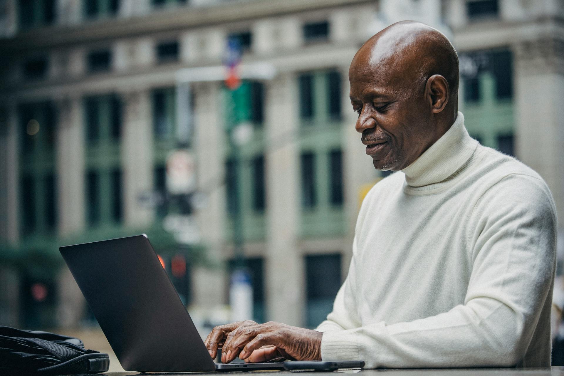 Side view of self employed concentrated mature bald black man in stylish turtleneck sitting in street cafe and typing on netbook during work