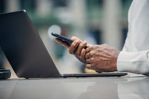 Crop anonymous elderly black businessman in casual outfit messaging on smartphone while sitting in street cafe during remote work on laptop