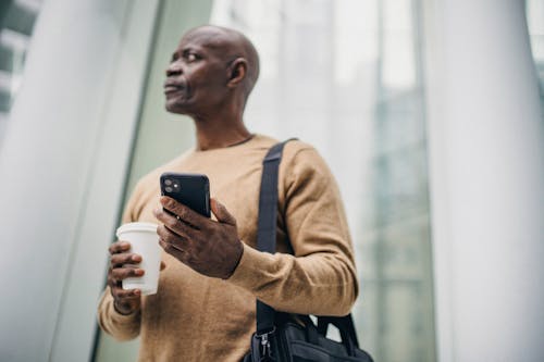Serious black man browsing phone while standing on street with takeaway coffee