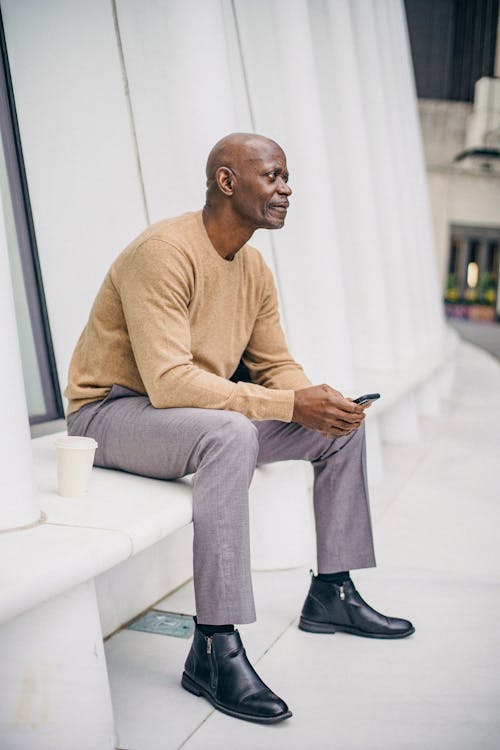 Free Side view of confident bald mature black male entrepreneur in stylish outfit browsing mobile phone while having coffee break on city bench Stock Photo