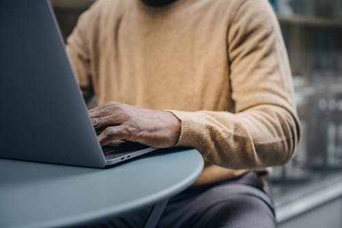 Free Crop unrecognizable mature ethnic male freelancer in stylish sweater typing on netbook keyboard while working remotely in outdoor cafe Stock Photo