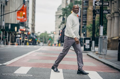 Side view of positive mature African American male pedestrian in elegant clothes smiling while crossing road in city downtown