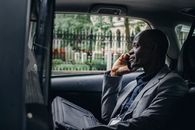 一位穿着优雅西装、成熟稳重的非裔美国商人，坐在出租车乘客座位上，正用智能手机通话的侧面图