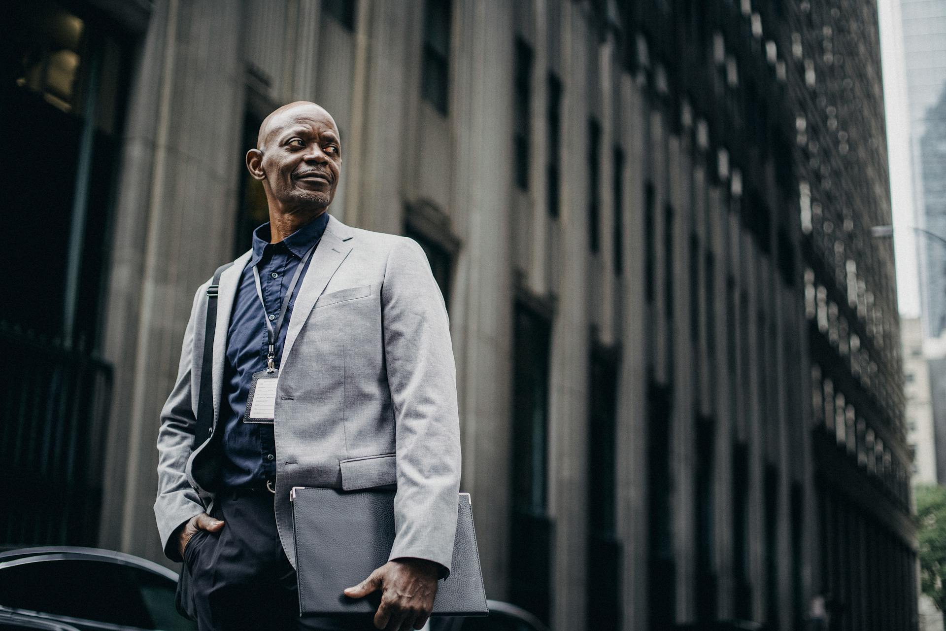 Low angle of confident bald black mature male employee in classy suit and badge standing on city street with folder in hand and looking away