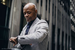 Serious African American businessman in formal suit looking at wristwatch while standing outside modern business center building