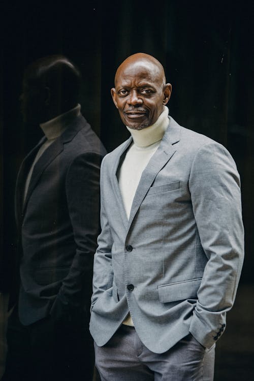 Free Positive adult African American businessman in classy formal suit standing with hands in pocket against black glass wall and looking at camera confidently Stock Photo