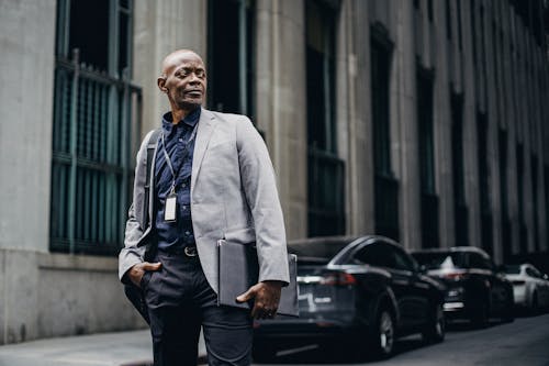 Serious adult African American businessman in formal suit standing with hand in pocket on urban street and looking away thoughtfully
