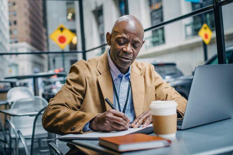 Black Male Office Worker Writing On Paper Near Laptop Indoors